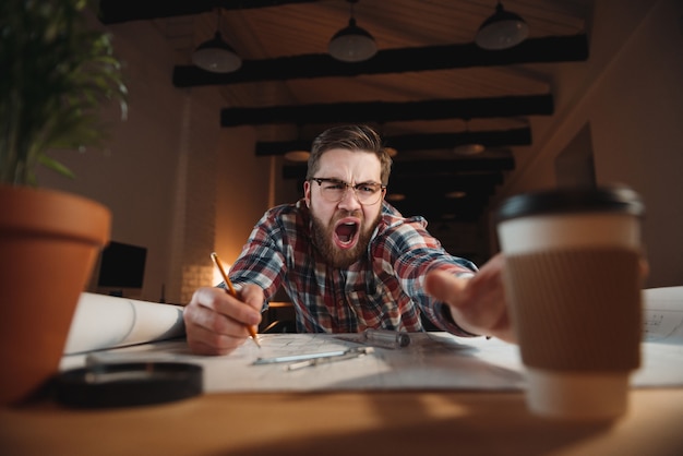 Portrait d'un travailleur barbu fatigué essayant d'atteindre une tasse de café et bâillant au bureau