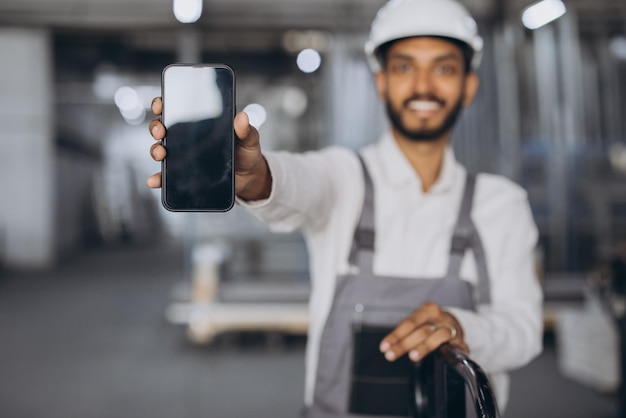 Portrait d'un travailleur hindou heureux en casque blanc et salopette tenant un smartphone et montrant un blanc