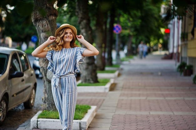 Portrait d'une très jolie jeune femme en combinaison rayée posant avec son chapeau sur un trottoir en ville