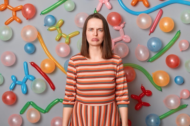 Portrait d'une triste femme désespérée aux cheveux bruns portant une robe rayée debout contre un mur gris avec des ballons colorés regardant la caméra avec tristesse personne ne vient à sa fête d'anniversaire