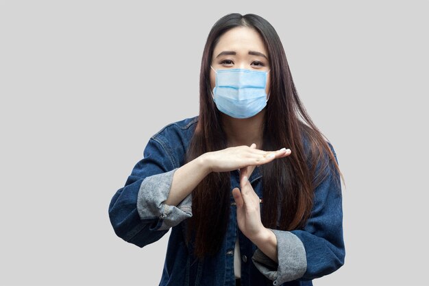 Portrait triste d'une jeune femme asiatique brune avec un masque médical chirurgical dans une veste en jean bleu décontractée debout et regardant la caméra avec un signe de délai d'attente. tourné en studio intérieur, isolé sur fond gris.