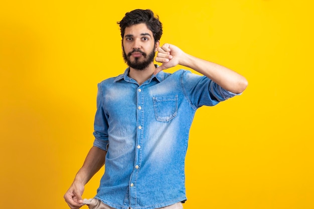 Portrait d'un triste jeune homme barbu en faillite en chemise bleue de style décontracté debout et montrant la poche vide et regardant la caméra prise de vue en studio à l'intérieur isolée sur jaune