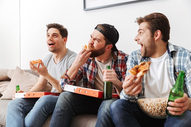 Portrait de trois hommes affamés de manger de la pizza et de boire de la bière avec, tout en soutenant l'équipe de football à la maison