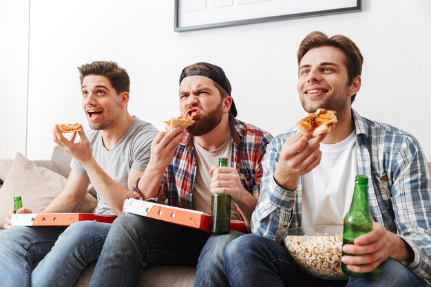 Portrait de trois jeunes célibataires mangeant de la pizza avec plaisir, tout en regardant un match de football à la télévision à la maison