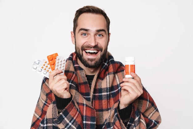 Portrait d'un type caucasien souriant enveloppé dans une couverture tenant un tas de comprimés à cause de la grippe isolée sur un mur blanc