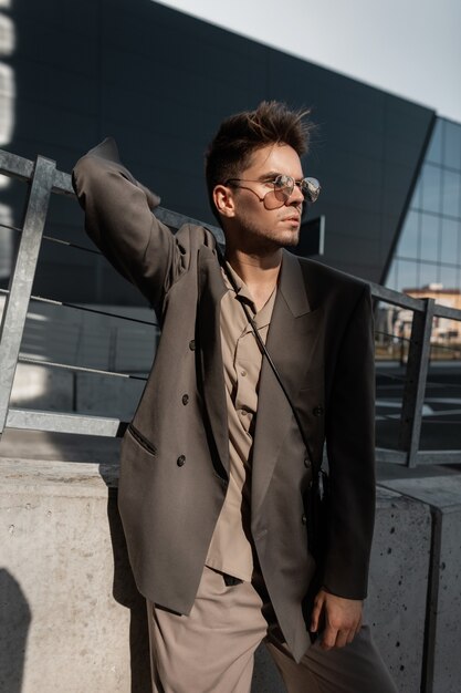 Portrait urbain élégant d'un beau jeune homme à poils dans un costume à la mode avec une chemise et un blazer dans des lunettes de soleil cool se dresse sur la ville au soleil