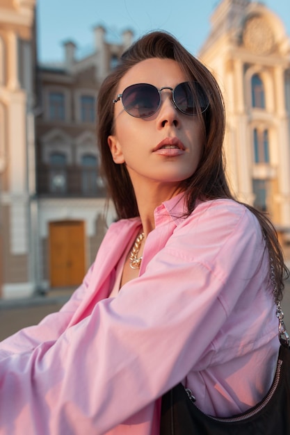 Photo portrait urbain à la mode d'une belle jeune femme avec des lunettes de soleil dans une chemise rose à la mode et un sac à main dans la ville