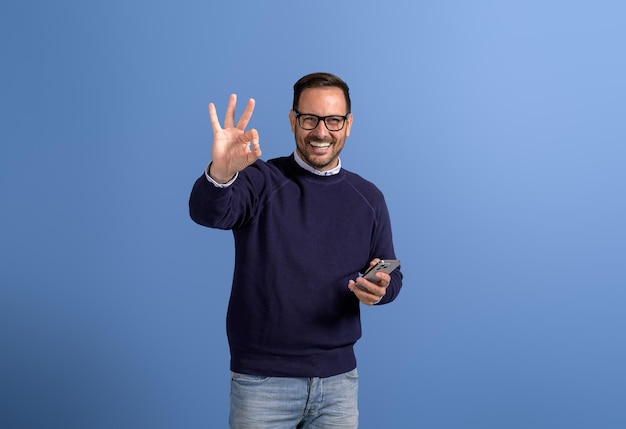 Photo portrait d'un vendeur souriant utilisant un téléphone portable et faisant des gestes signe ok sur fond bleu isolé