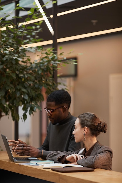 Portrait vertical aux tons chauds de deux jeunes collègues discutant du travail au bureau debout dans un bureau moderne