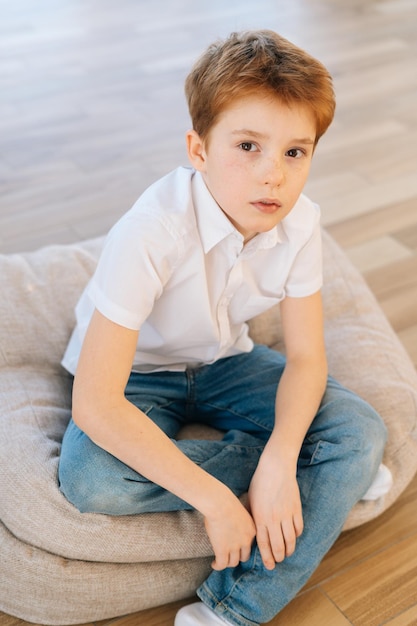 Portrait vertical d'un bel enfant garçon regardant la caméra assis sur un oreiller au sol dans le salon avec un intérieur moderne et lumineux