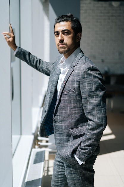 Portrait vertical d'un bel homme d'affaires portant un costume de mode debout près de la fenêtre dans une salle de bureau moderne regardant la caméra