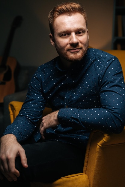 Portrait vertical d'un bel homme barbu portant une chemise élégante assis confortablement dans un luxueux fauteuil jaune