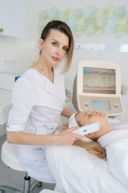 Portrait vertical d'une cosmétologue effectuant un ultrasound, un massage de lifting du visage avec un équipement professionnel pour une jeune femme dans une clinique de médecine esthétique.