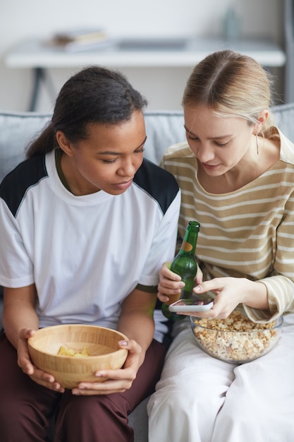 Portrait vertical de deux jeunes femmes regardant la télévision à la maison et mangeant du pop-corn