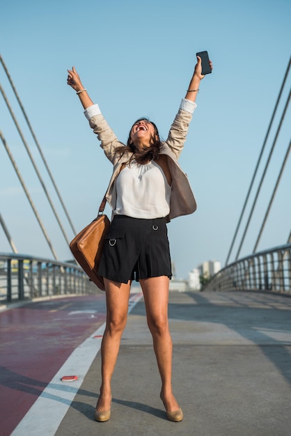 Portrait vertical d'une femme d'affaires levant les bras en triomphe avec un téléphone portable à la main et une mallette dans la rue Espace de copie