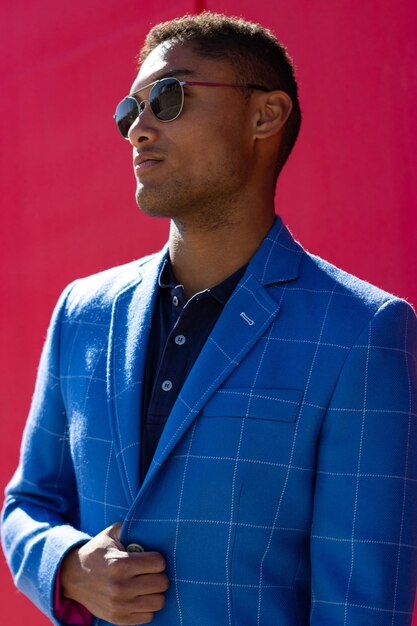 Portrait vertical d'un homme d'affaires noir latin avec lunettes de soleil et blazer bleu sur fond rouge