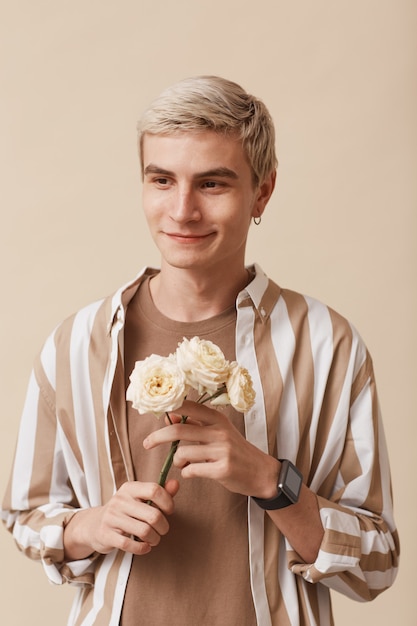 Portrait vertical d'un homme blond féminin tenant des fleurs et souriant en se tenant debout sur un fond beige neutre en studio