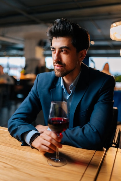 Portrait vertical d'un homme élégant réussi en costume de mode tenant des verres de vin rouge assis à table dans un restaurant à la recherche de suite