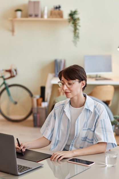 Portrait vertical de jeune femme à l'aide d'une tablette à stylet sur le lieu de travail du bureau à domicile pour la conception numérique ou pho