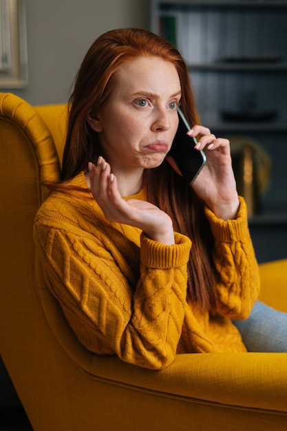 Portrait vertical d'une jeune femme déconcertée parlant sur un smartphone assis sur une chaise jaune à la maison Jolie dame choquée ayant une conversation parlant par téléphone