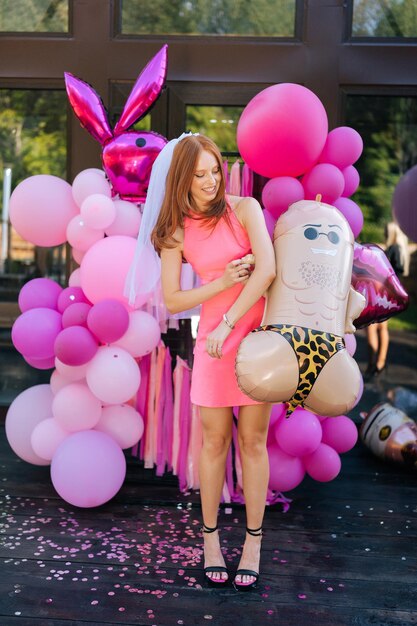 Portrait vertical d'une jeune femme jolie et joyeuse en robe d'été posant tenant des ballons d'hélium en papier d'aluminium festif coloré drôle debout sur le fond d'une maison de campagne regardant loin avec une expression heureuse