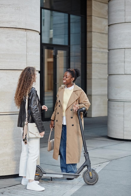 Portrait vertical d'une jeune femme noire avec un scooter électrique parlant à un ami en ville