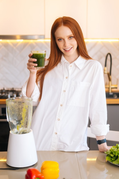 Portrait vertical d'une jeune femme rousse heureuse tenant un verre avec un smoothie de désintoxication aux légumes verts