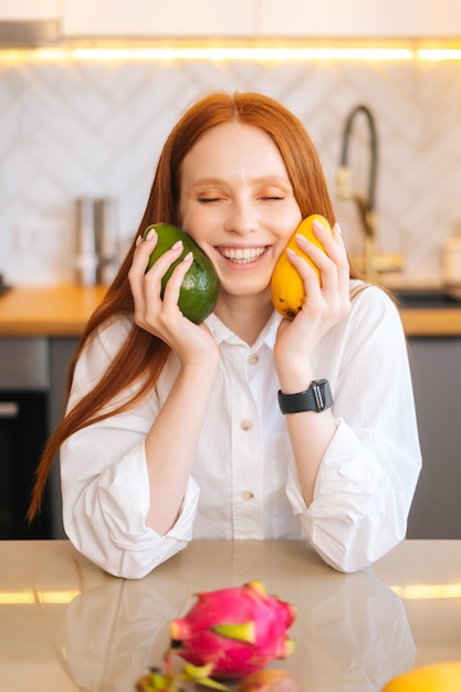 Portrait vertical d'une jeune femme rousse séduisante et joyeuse tenant dans les mains la mangue et l'avocat