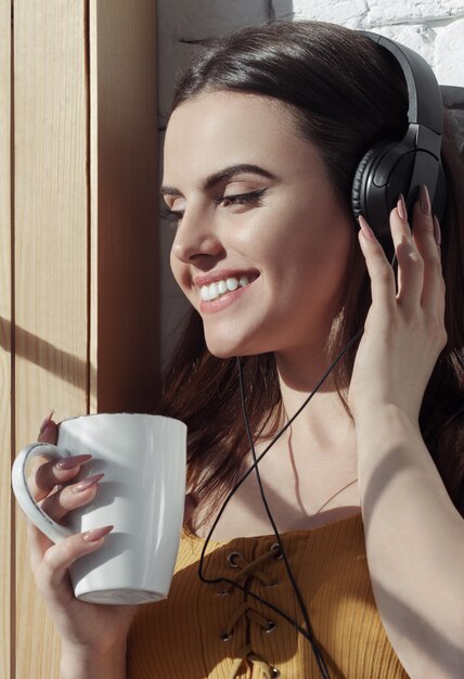 Portrait vertical de jeune fille souriante, écouter de la musique et boire du café sur le rebord de la fenêtre