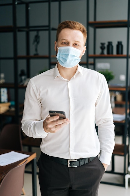 Portrait vertical d'un jeune homme d'affaires portant un masque protecteur à l'aide d'un téléphone portable debout dans une salle de bureau moderne, regardant la caméra. Employé confiant utilisant un smartphone pour planifier son temps de travail.