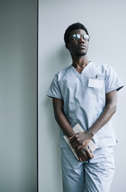 Photo portrait vertical d'un jeune médecin afro-américain debout près du mur dans une clinique et regardant loin