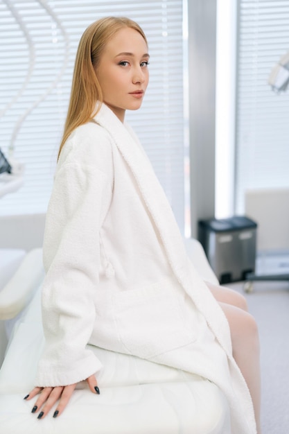 Photo portrait vertical d'une jolie patiente en robe de bain blanche assise sur un canapé médical moderne