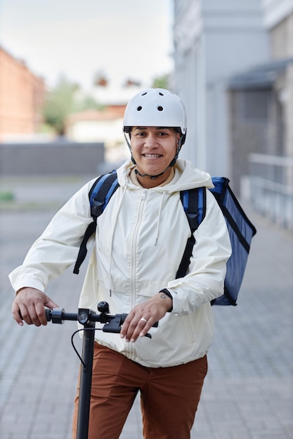 Portrait vertical d'un livreur de nourriture souriant portant un casque tout en faisant du scooter électrique en ci