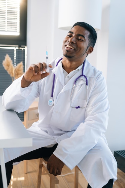 Portrait vertical d'un médecin afro-américain joyeux portant un uniforme blanc tenant une seringue à la main assis à table dans une clinique médicale