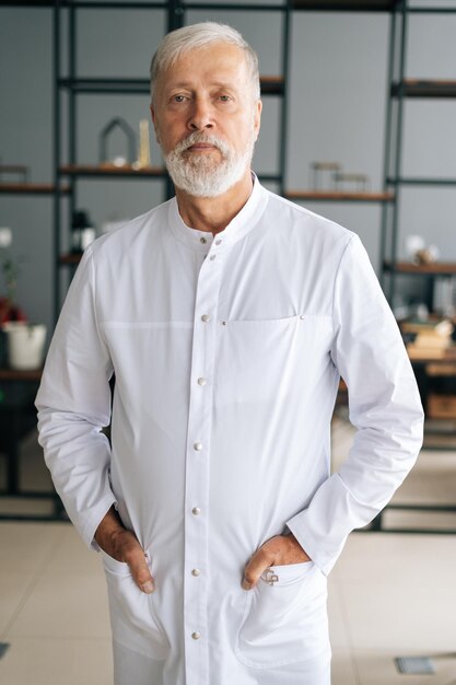 Portrait vertical d'un médecin de sexe masculin adulte senior sérieux en uniforme médical blanc debout dans le bureau de l'hôpital regardant la caméra