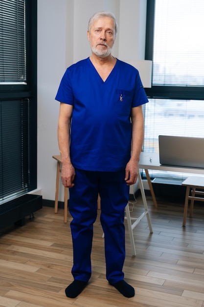 Portrait vertical de pleine longueur d'un médecin de sexe masculin adulte mature fatigué en uniforme médical debout dans le bureau de l'hôpital sur fond de fenêtre regardant la caméra