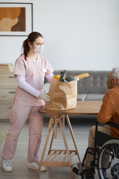 Portrait vertical de toute la longueur d'une jeune infirmière apportant des provisions à un homme âgé en fauteuil roulant, concept d'assistance et de livraison de nourriture