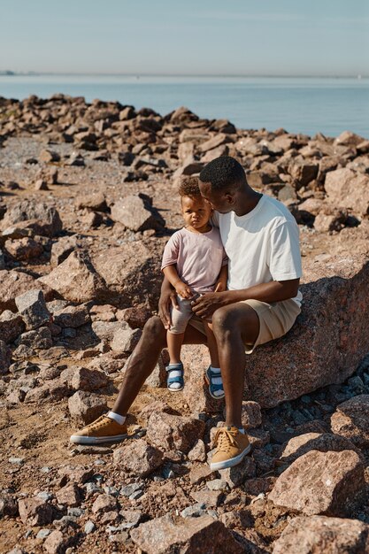 Portrait vertical de toute la longueur d'un jeune père afro-américain jouant avec un fils mignon sur la plage