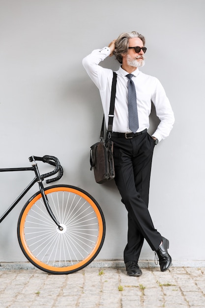 Portrait d'un vieil homme d'affaires respectable en lunettes de soleil regardant de côté en se tenant debout avec un vélo sur un mur gris à l'extérieur