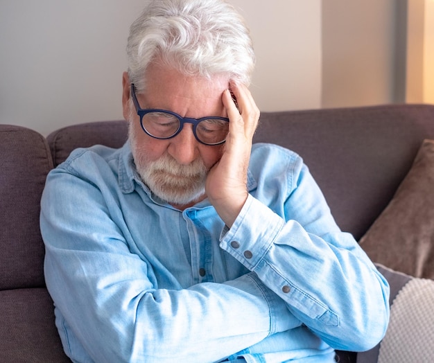 Portrait d'un vieil homme malheureux et triste aux cheveux blancs assis sur le canapé à la maison avec la main sur le visage déprimé