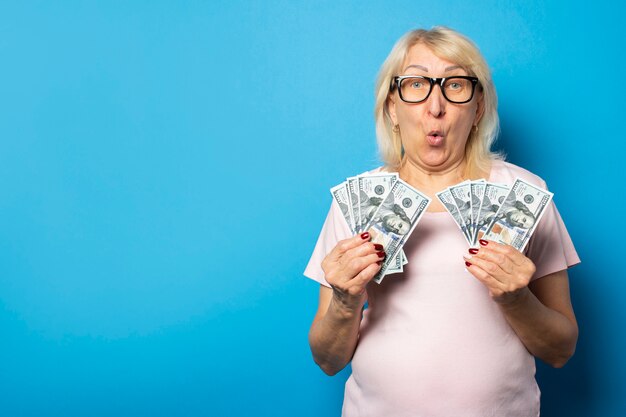 Portrait d'une vieille femme sympathique avec un visage surpris dans un t-shirt décontracté et des lunettes tenant de l'argent dans ses mains sur un mur bleu isolé. Visage émotionnel. Concept richesse, victoire, prêt, pension