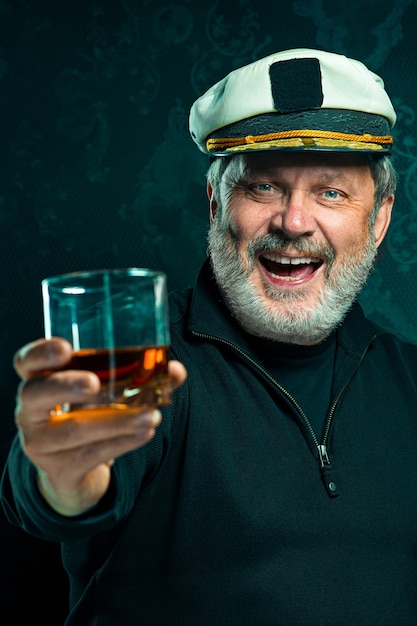Portrait d'un vieux marin en tant que capitaine en pull noir et chapeau buvant du cognac sur fond de studio noir