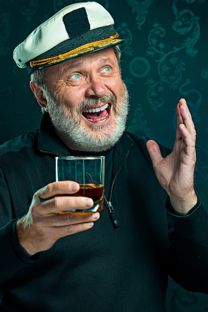 Portrait d'un vieux marin en tant que capitaine en pull noir et chapeau buvant du cognac sur fond de studio noir