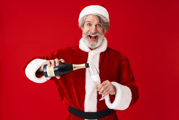 Portrait d'un vieux père Noël aux cheveux gris heureux souriant et tenant une bouteille de champagne isolée sur rouge en studio