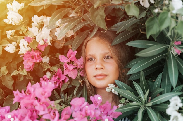 Portrait visage d'une belle petite fille candide de huit ans aux yeux bruns sur fond de plantes vertes et de fleurs roses pendant des vacances d'été voyage gen z concept de santé mentale flare