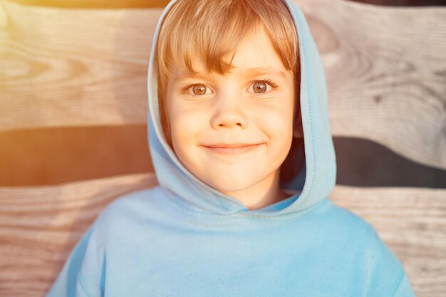 Portrait de visage heureux souriant franc garçon de cinq ans en sweat à capuche bleu dans une capuche sur le fond de planches de bois dans la nature de l'été dans une heure d'or au coucher du soleil homeschool et travel flare