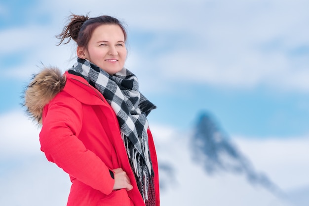 Portrait de voyageuse vêtue d'une veste coupe-vent d'hiver rouge, d'une écharpe noire et blanche autour du cou. Mystérieuse belle jeune femme sur fond de montagnes et de ciel bleu avec des nuages blancs.