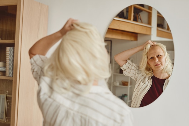 Portrait de vue arrière aux tons chauds de femme chauve décollant la perruque tout en regardant dans le miroir à l'intérieur de la maison, l'alopécie et la sensibilisation au cancer, copiez l'espace