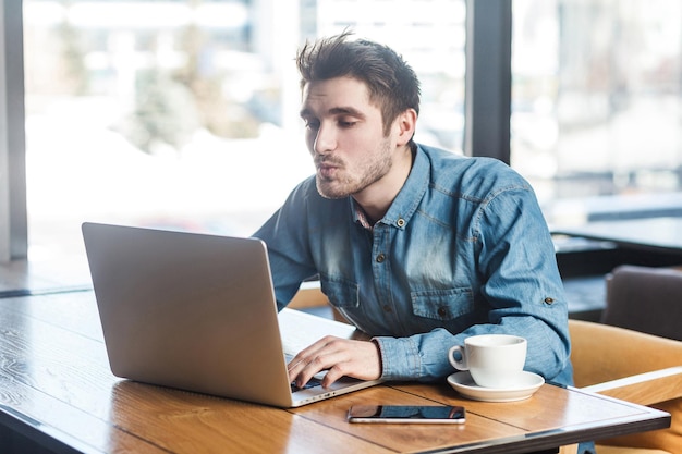 Portrait de vue latérale d'un beau jeune homme barbu en chemise de jeans bleu assis seul dans un café et faisant un appel vidéo en envoyant un baiser aérien à sa petite amie sur un ordinateur portable.