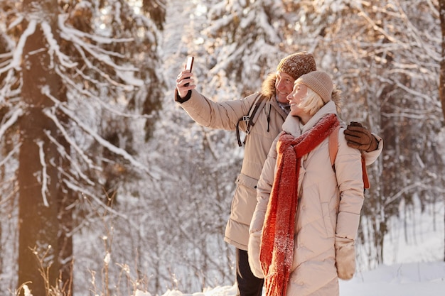 Portrait de vue latérale d'un couple mature actif prenant une photo de selfie tout en profitant d'une randonnée dans la forêt d'hiver